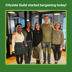 Four people stand in an office smiling with their arms around each other