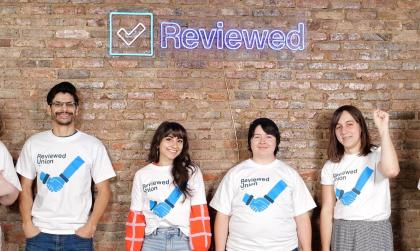 Four people wearing white Reviewed union t-shirts stand against a brick wall, smiling